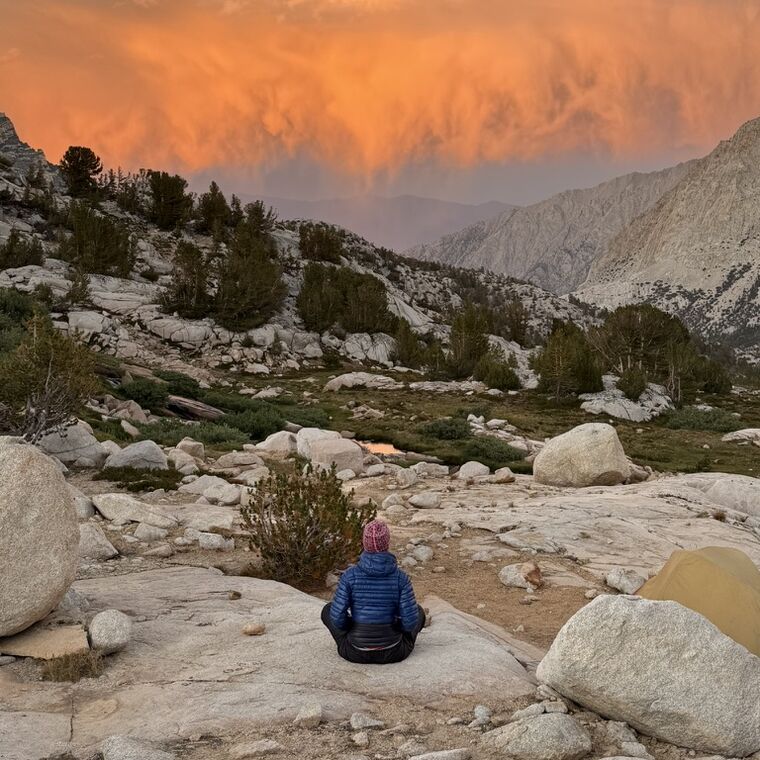 A hiker sitting on granite, watching a wall of cloud turn orange over the ridge