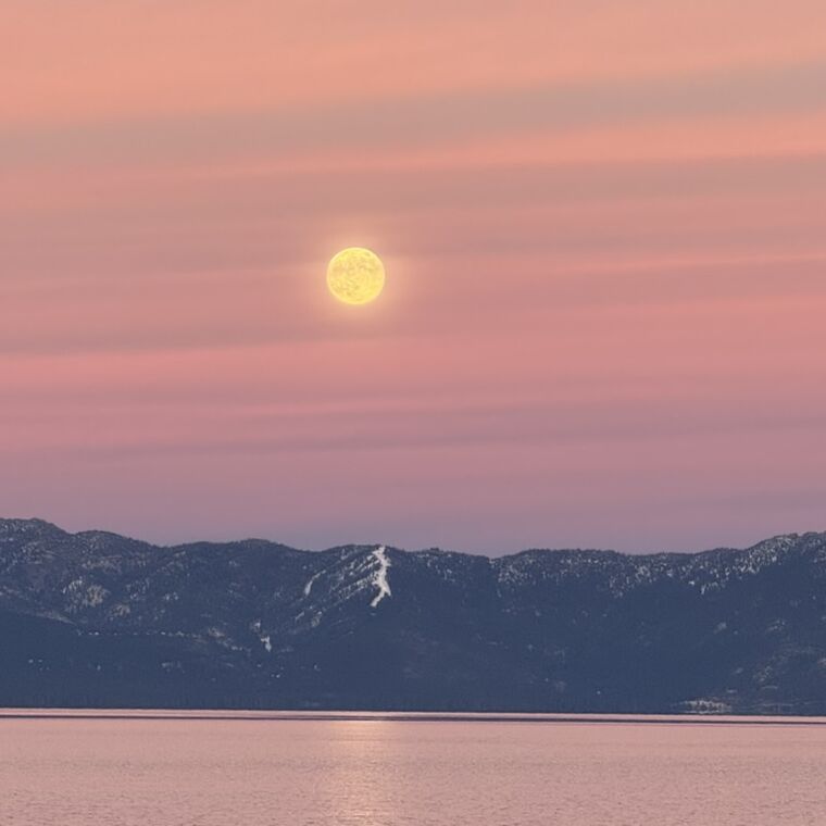 A full moon rising into a pink sky above the snow-streaked ridge across the lake