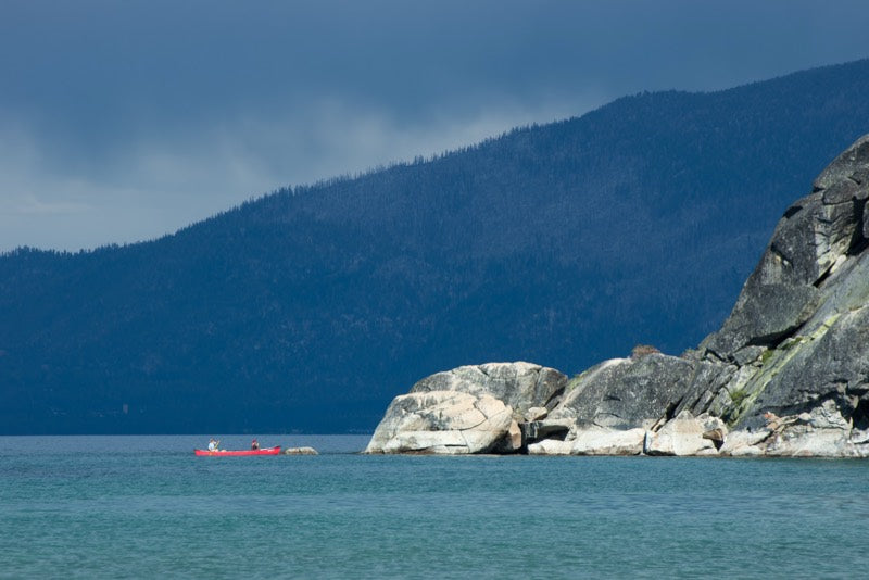 A single red canoe crossing Lake Tahoe below a dark forested ridge