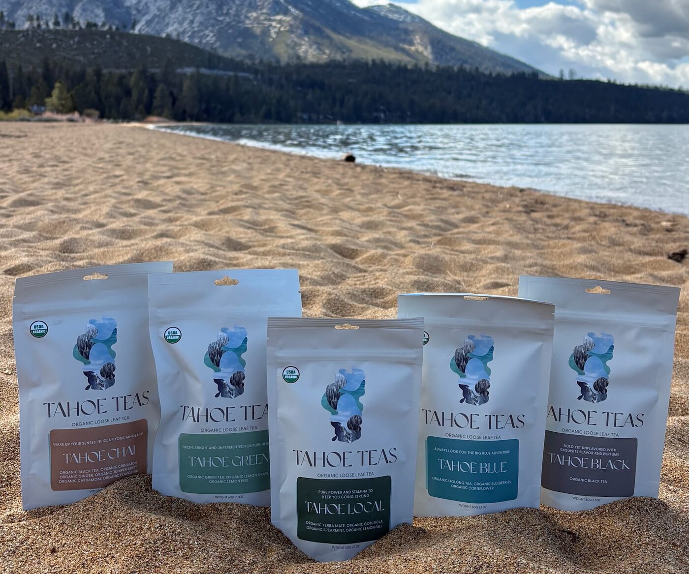 Five Tahoe Teas pouches standing in the sand on a Lake Tahoe beach, the lake and Mount Tallac behind them