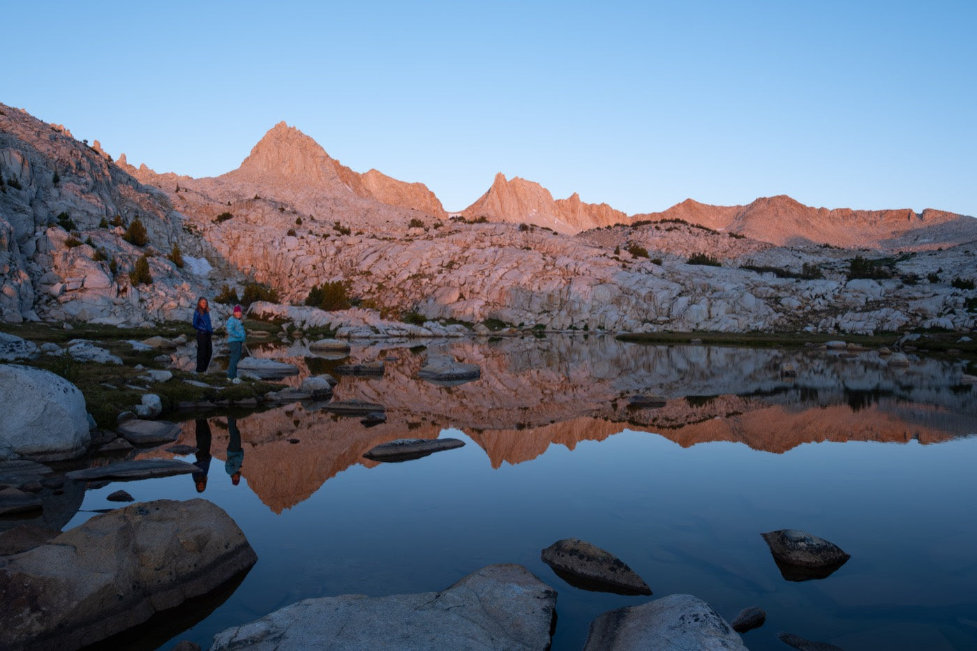 Two people at the edge of a High Sierra lake at first light, the peaks reflected in still water